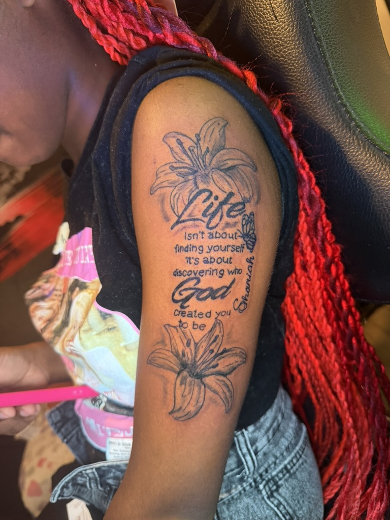 Blackwork upper-arm tattoo with lilies, butterfly, and cursive script